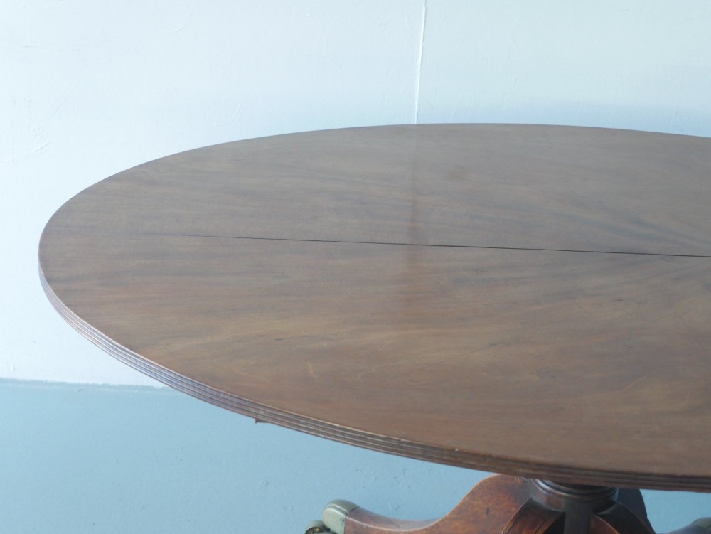 An antique Regency oval mahogany tilt-top table shown upright on a splayed pedestal base with brass castors. View V