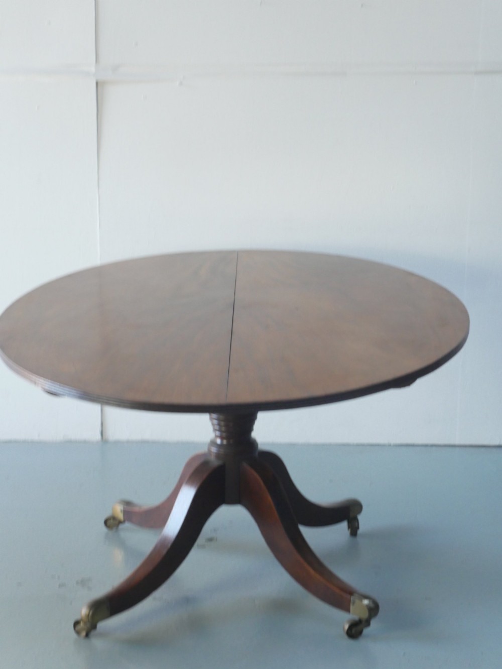 An antique Regency oval mahogany tilt-top table shown upright on a splayed pedestal base with brass castors. View VII