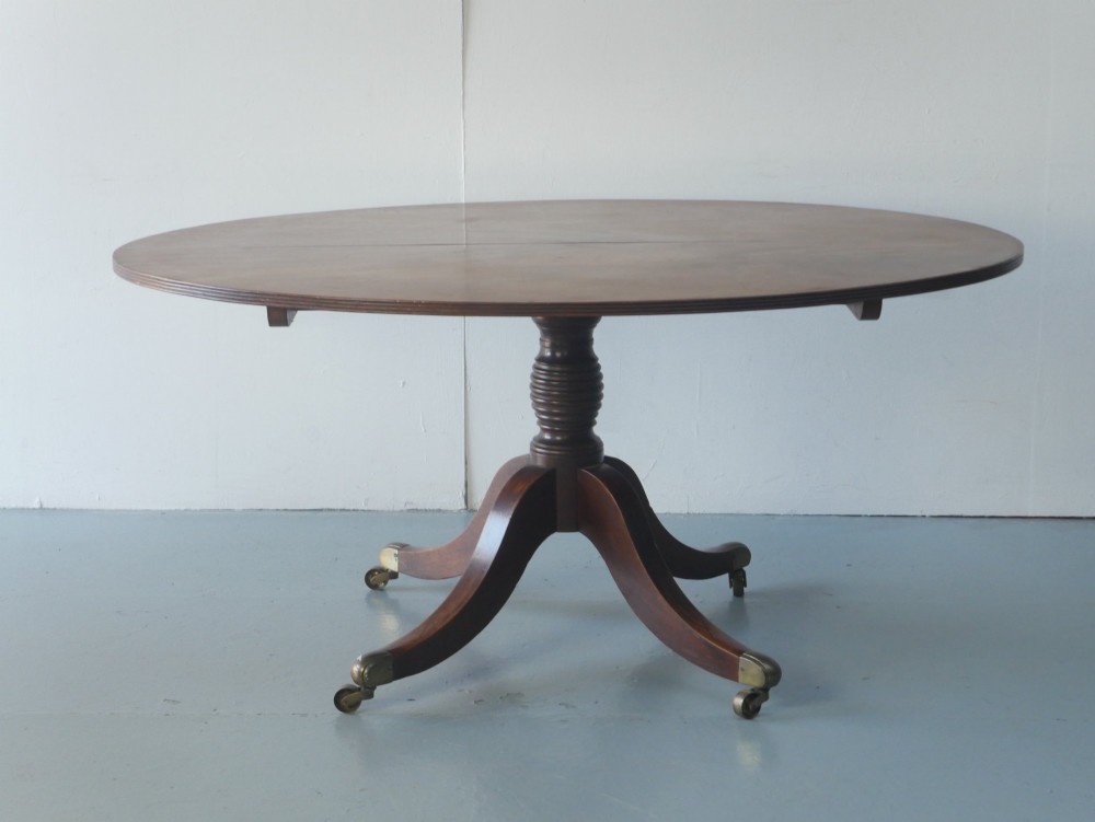 An antique Regency oval mahogany tilt-top table shown upright on a splayed pedestal base with brass castors. View X