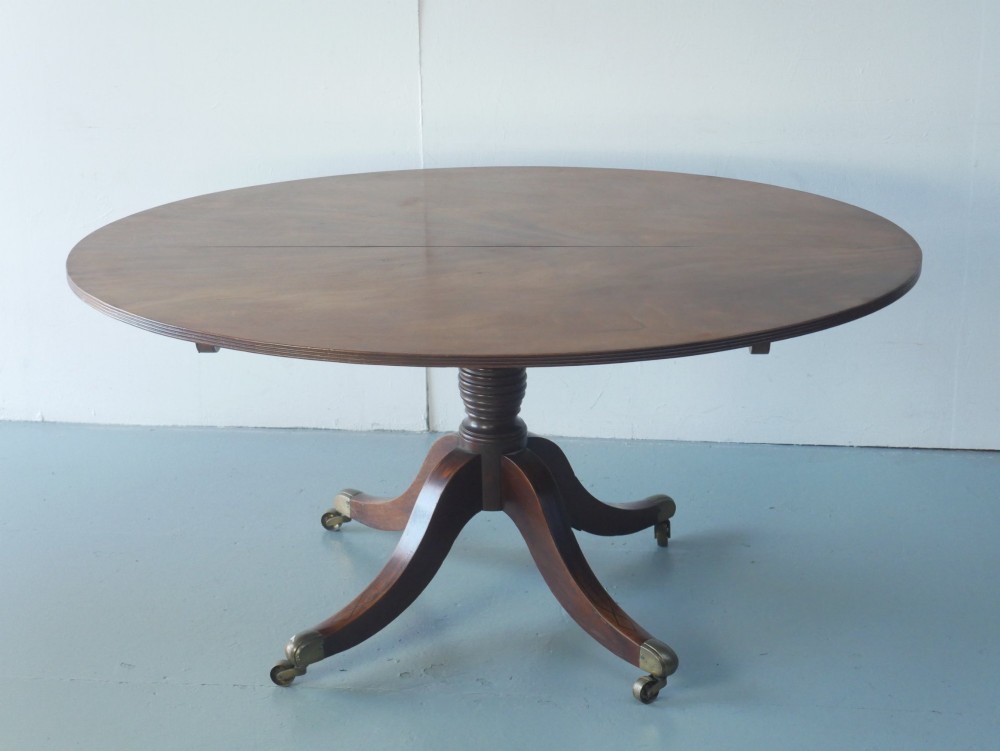 An antique Regency oval mahogany tilt-top table shown upright on a splayed pedestal base with brass castors. View XI
