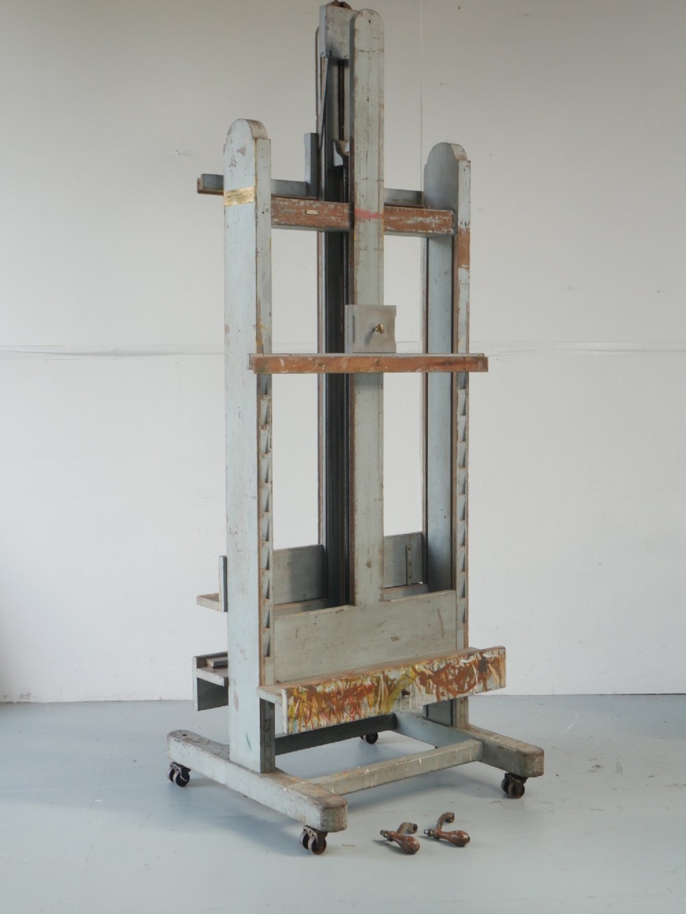 A large floor-standing double-sided timber studio easel with a distressed grey finish, adjustable height mechanism, and metal castor feet.