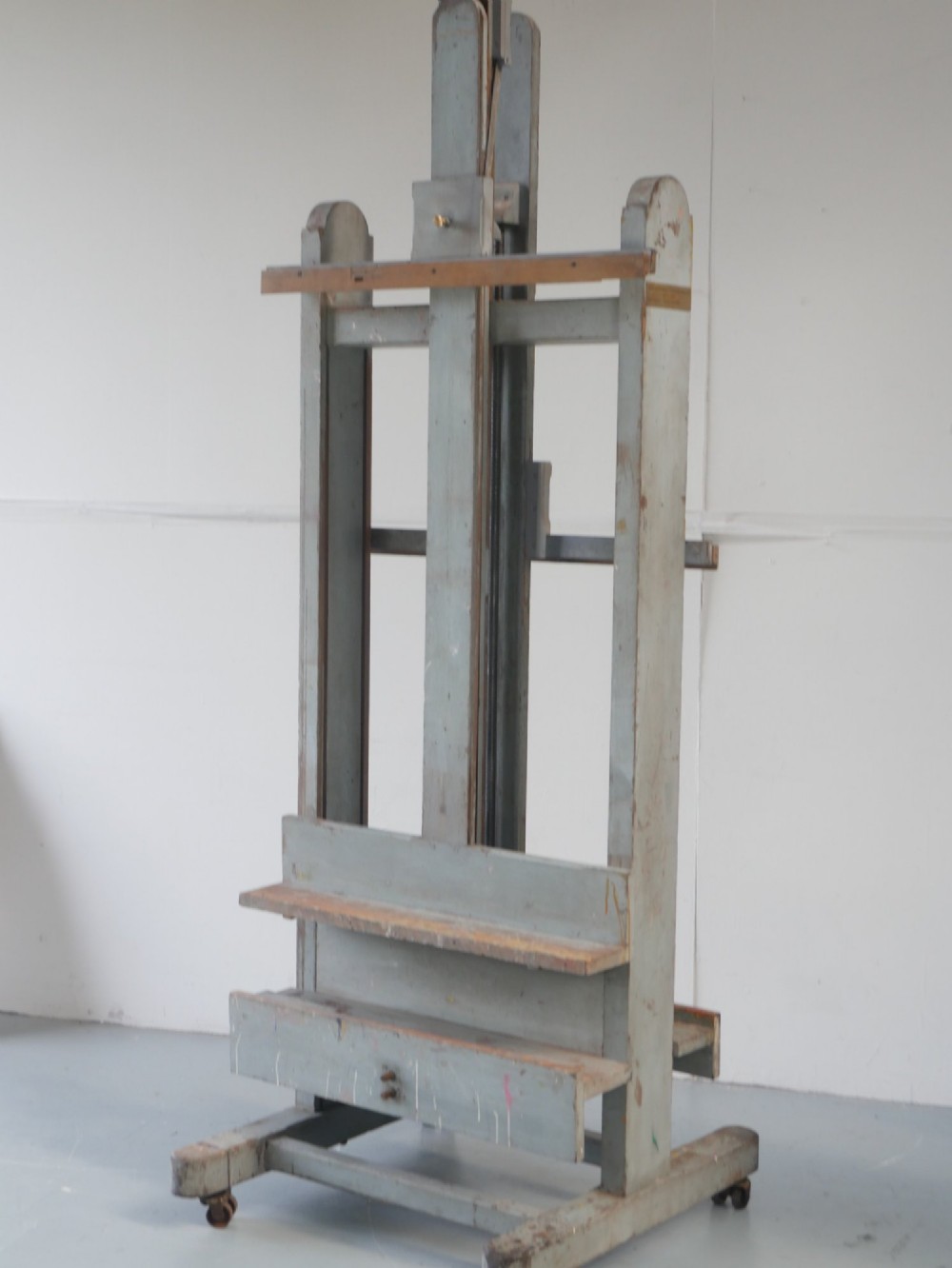 A large floor-standing double-sided timber studio easel with a distressed grey finish, adjustable height mechanism, and metal castor feet. View III
