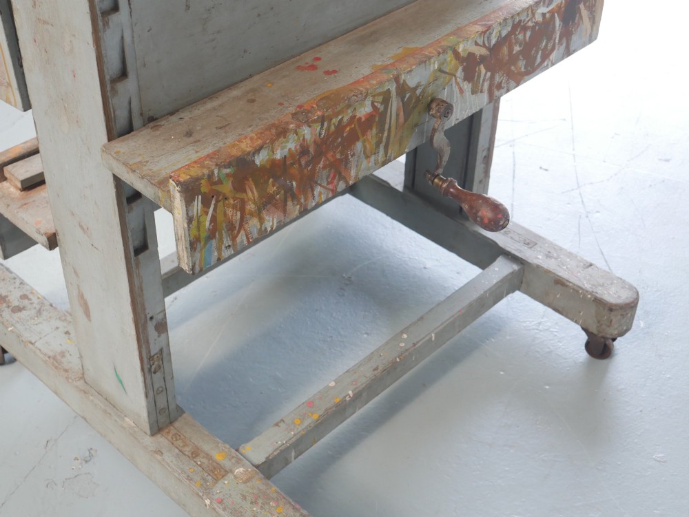 A large floor-standing double-sided timber studio easel with a distressed grey finish, adjustable height mechanism, and metal castor feet. View V