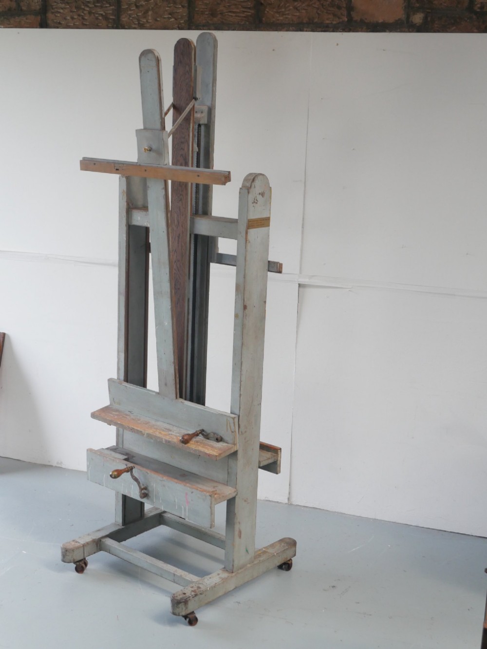A large floor-standing double-sided timber studio easel with a distressed grey finish, adjustable height mechanism, and metal castor feet. View VI