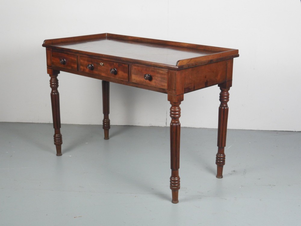 A Regency period mahogany writing table featuring three drawers with wooden knobs, a three-sided gallery rim, and four reeded legs.