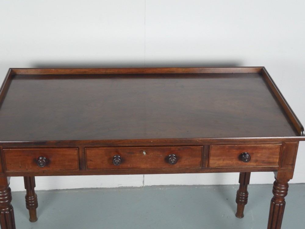 A Regency period mahogany writing table featuring three drawers with wooden knobs, a three-sided gallery rim, and four reeded legs. View II