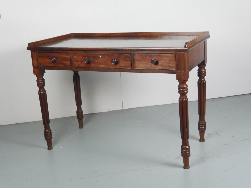 A Regency period mahogany writing table featuring three drawers with wooden knobs, a three-sided gallery rim, and four reeded legs. View IV