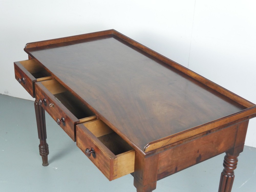 A Regency period mahogany writing table featuring three drawers with wooden knobs, a three-sided gallery rim, and four reeded legs. View VIII