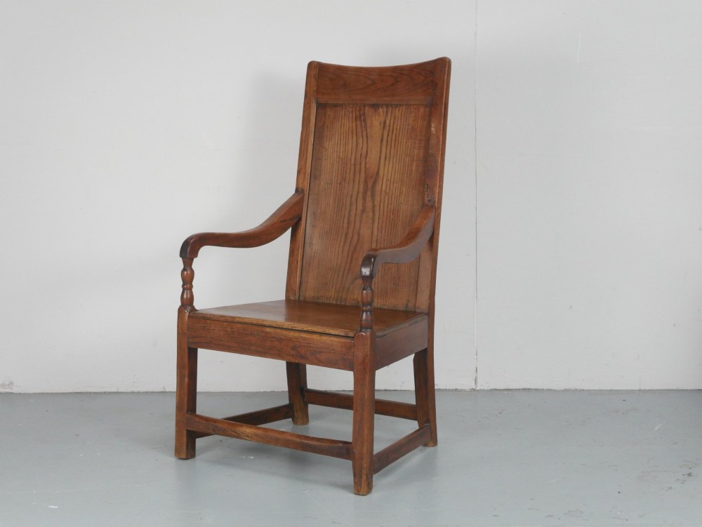 An early 19th-century country oak armchair featuring a high panelled backrest, curved arms, turned front supports, and a square stretcher base.