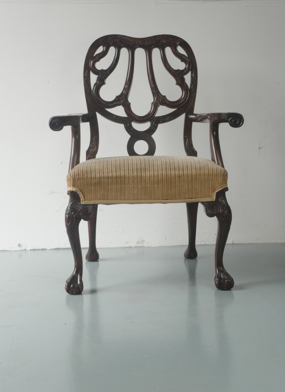 A carved mahogany armchair featuring an ornate pierced splat back, scrolled arms, tan striped fabric upholstery, and cabriole front legs with claw-and-ball feet.