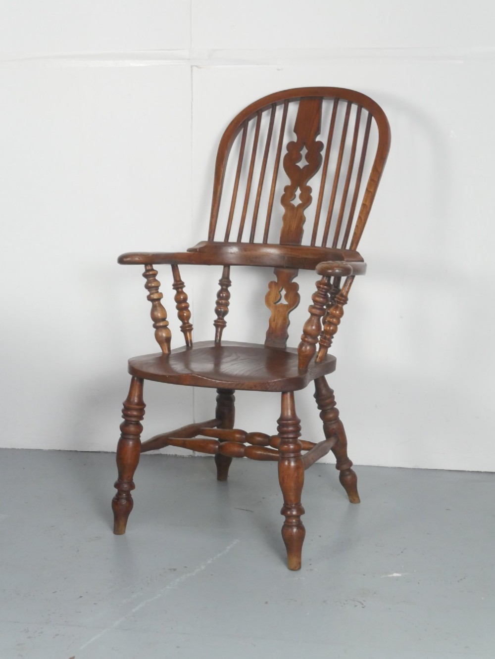 An antique wooden Windsor armchair with a hoop back, central pierced splat, spindle backrest, broad armrests, and turned legs with an H-stretcher in a warm brown timber finish.