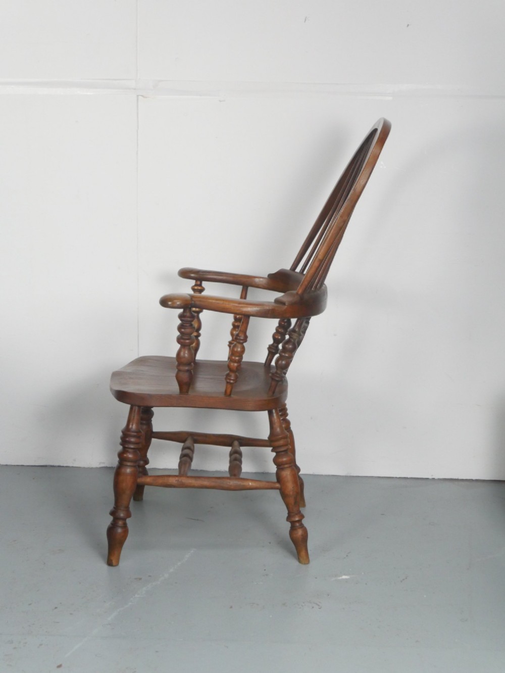 An antique wooden Windsor armchair with a hoop back, central pierced splat, spindle backrest, broad armrests, and turned legs with an H-stretcher in a warm brown timber finish. View II
