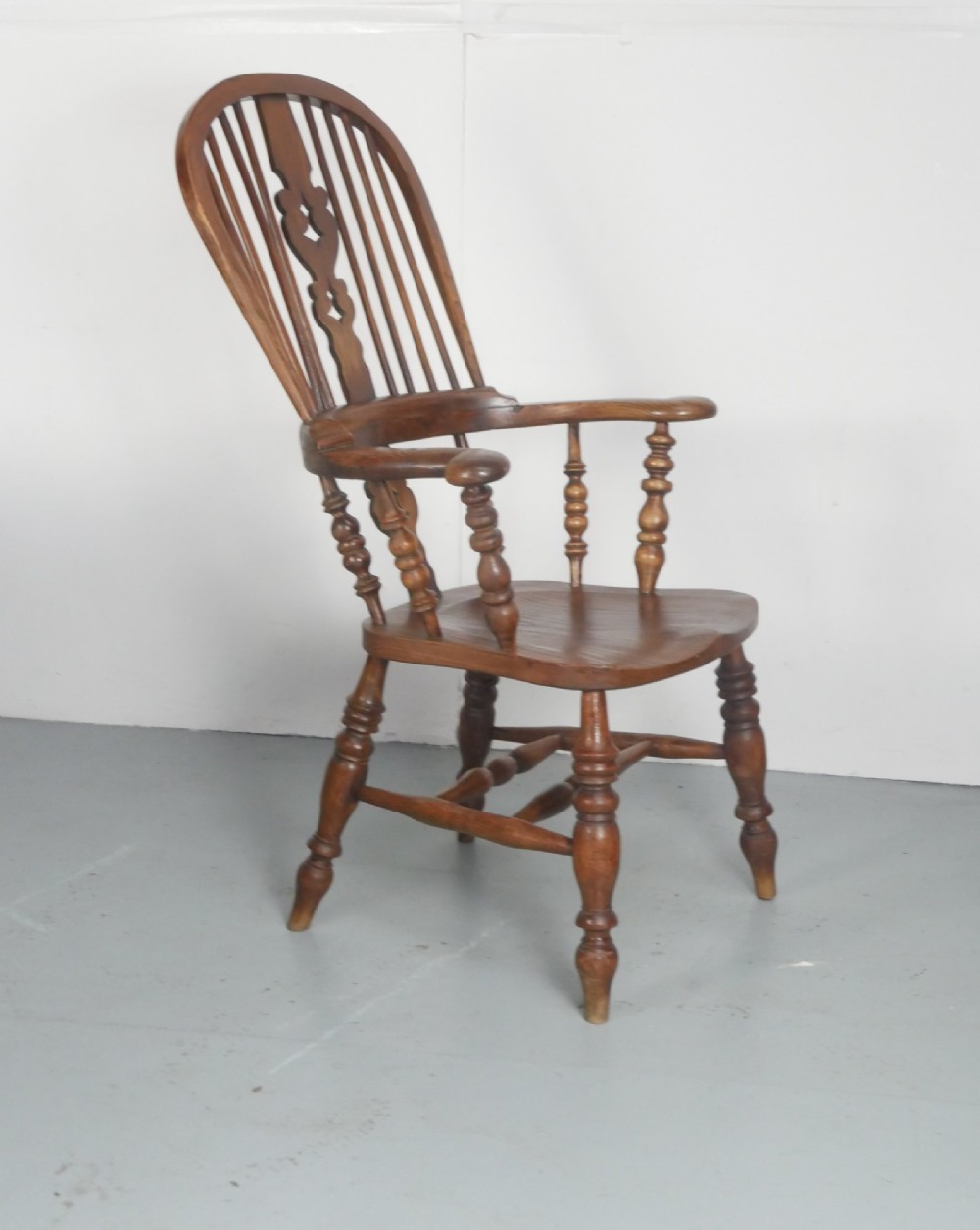 An antique wooden Windsor armchair with a hoop back, central pierced splat, spindle backrest, broad armrests, and turned legs with an H-stretcher in a warm brown timber finish. View III