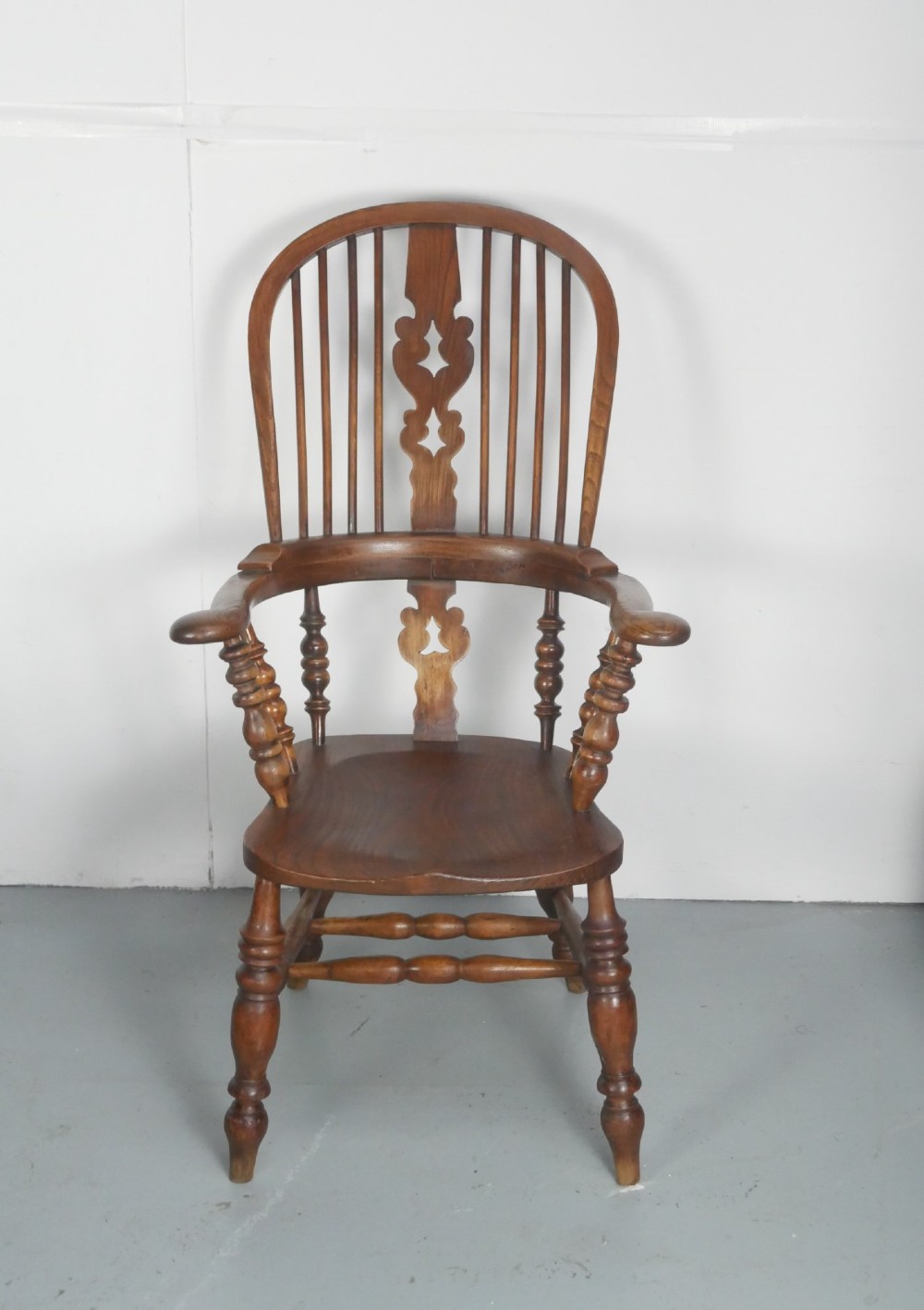 An antique wooden Windsor armchair with a hoop back, central pierced splat, spindle backrest, broad armrests, and turned legs with an H-stretcher in a warm brown timber finish. View IV