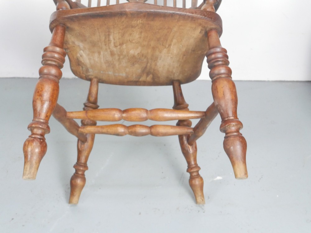 An antique wooden Windsor armchair with a hoop back, central pierced splat, spindle backrest, broad armrests, and turned legs with an H-stretcher in a warm brown timber finish. View V