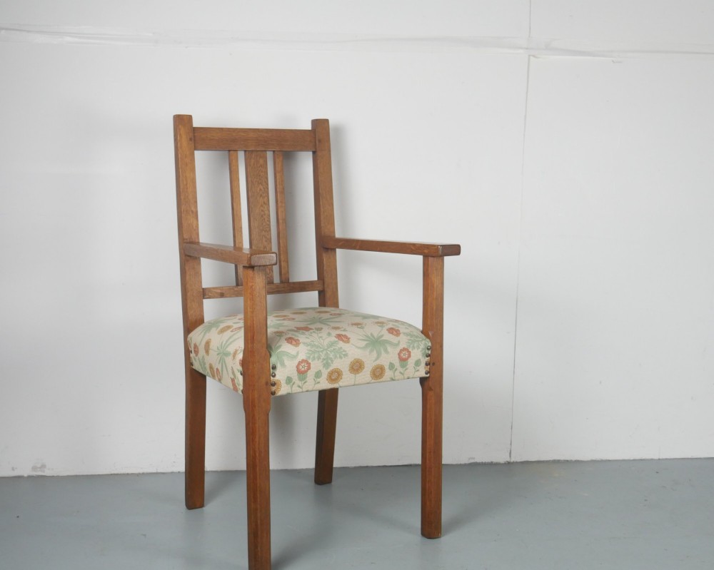 An Arts and Crafts style medium oak armchair featuring vertical back slats, flat armrests, and floral pattern upholstery with brass stud trim.