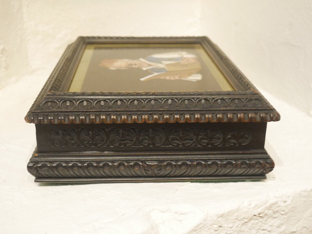 A rectangular dark carved wooden box featuring a detailed pietra dura hardstone inlay depicting an elderly man in spectacles reading a book. View V