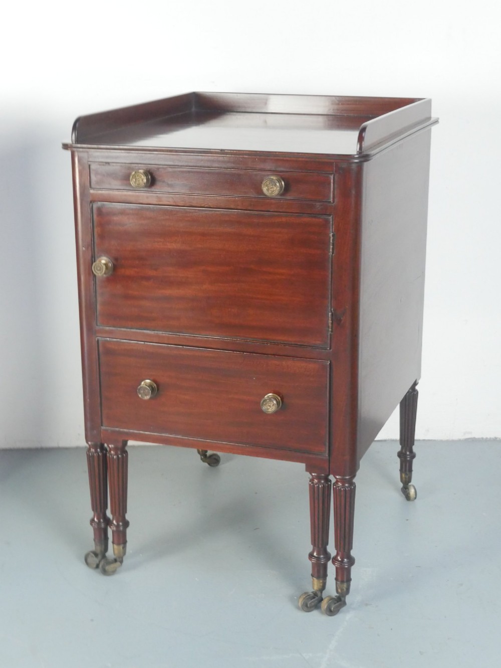 A matched pair of dark mahogany Regency bedside cupboards featuring gallery tops, single cupboard doors, lower drawers, reeded legs, and brass castors. View IX
