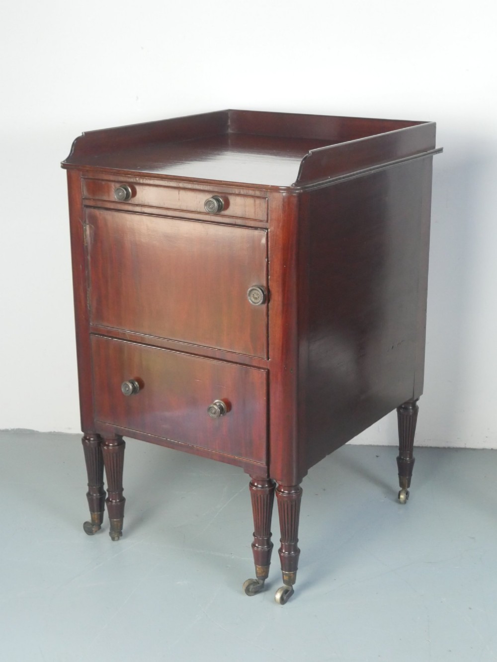 A matched pair of dark mahogany Regency bedside cupboards featuring gallery tops, single cupboard doors, lower drawers, reeded legs, and brass castors. View X