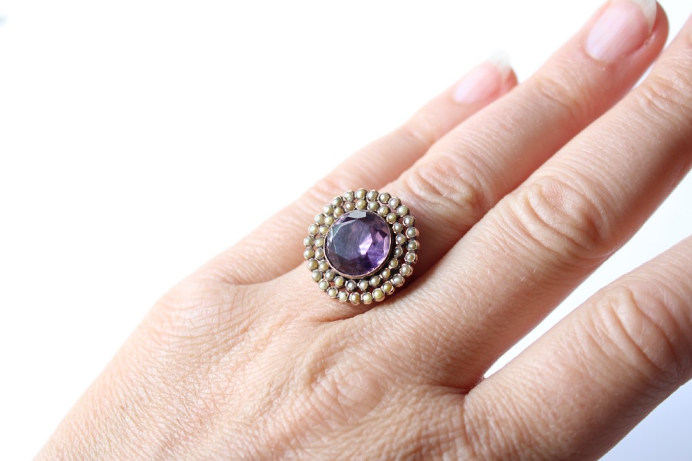 An antique 9ct yellow gold ring featuring a central oval purple amethyst stone surrounded by a double halo of cream seed pearls. View V