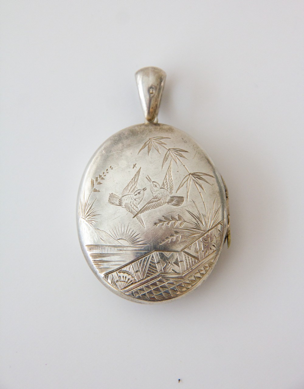 An oval silver locket featuring hand-engraved decoration of two birds, bamboo leaves, a sunburst motif, and geometric patterns. View II