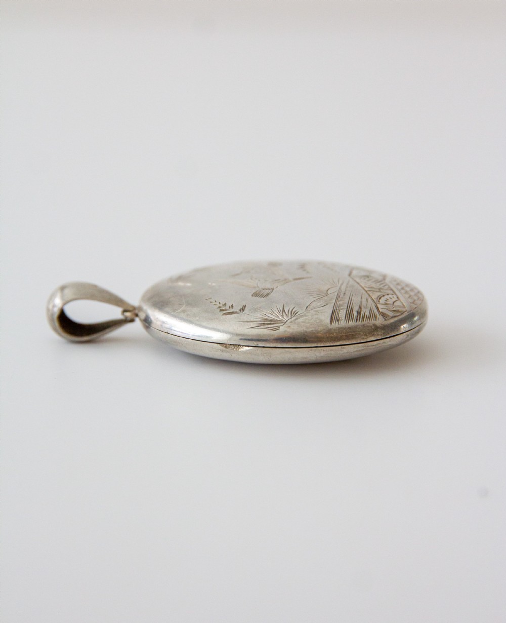 An oval silver locket featuring hand-engraved decoration of two birds, bamboo leaves, a sunburst motif, and geometric patterns. View III