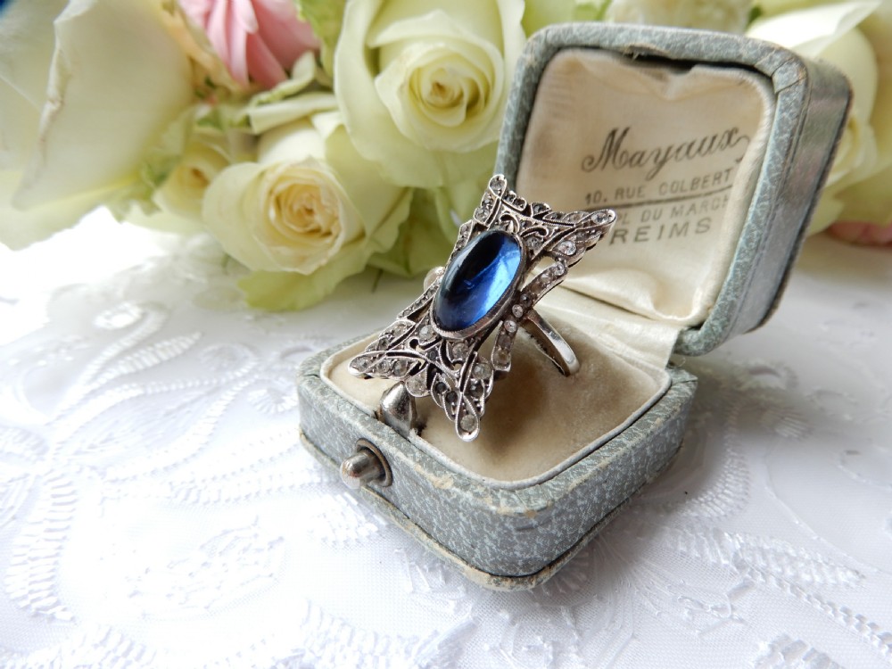 An Art Deco silver navette ring featuring an oval blue paste cabochon surrounded by clear paste stones in a geometric filigree setting.