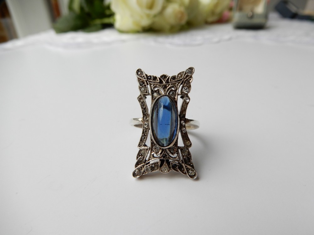 An Art Deco silver navette ring featuring an oval blue paste cabochon surrounded by clear paste stones in a geometric filigree setting. View II