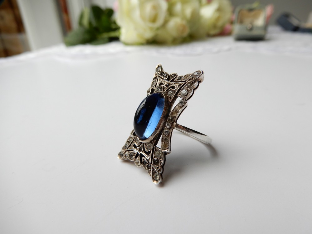 An Art Deco silver navette ring featuring an oval blue paste cabochon surrounded by clear paste stones in a geometric filigree setting. View III