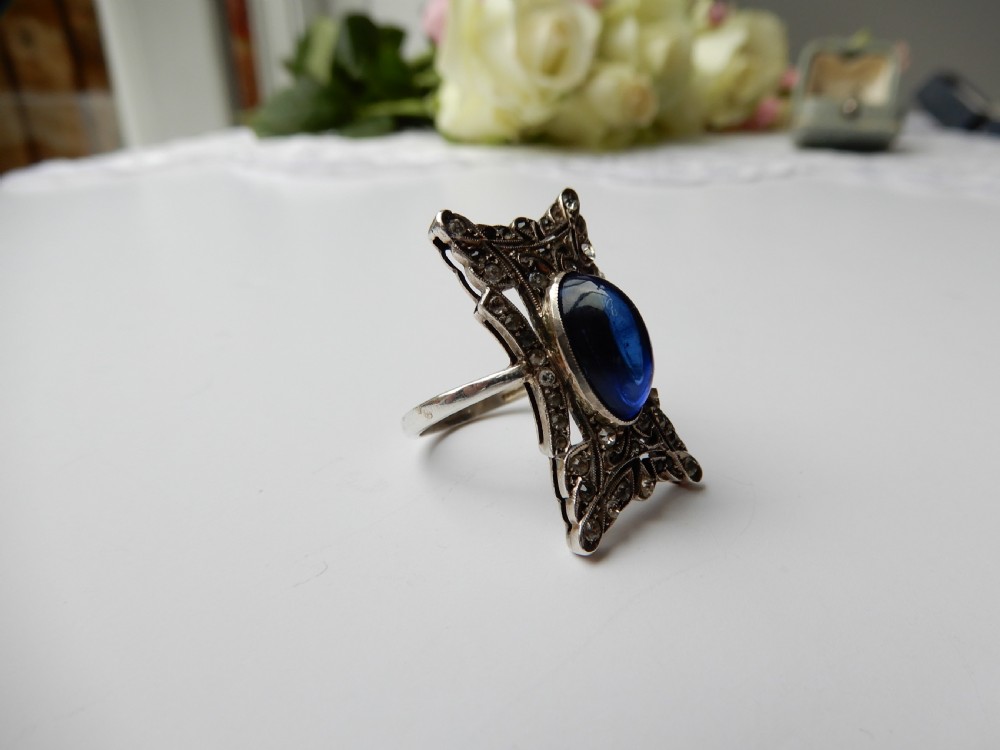 An Art Deco silver navette ring featuring an oval blue paste cabochon surrounded by clear paste stones in a geometric filigree setting. View IV