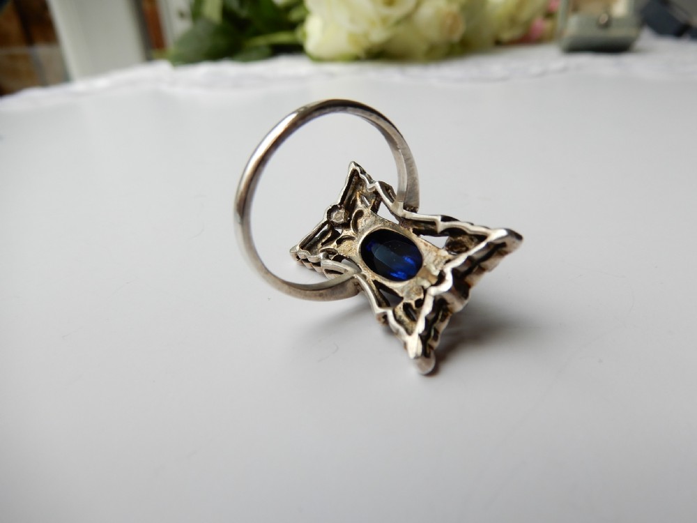An Art Deco silver navette ring featuring an oval blue paste cabochon surrounded by clear paste stones in a geometric filigree setting. View V