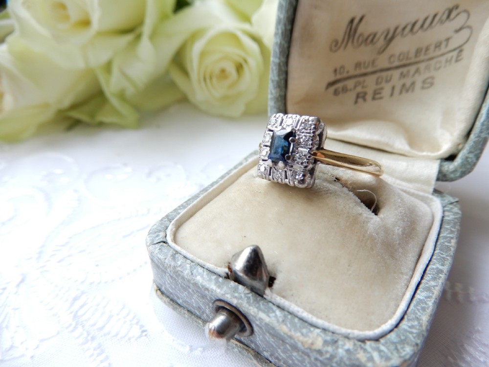 An Art Deco ring featuring a central rectangular blue sapphire surrounded by a geometric halo of small diamonds set in platinum atop an 18ct yellow gold band.