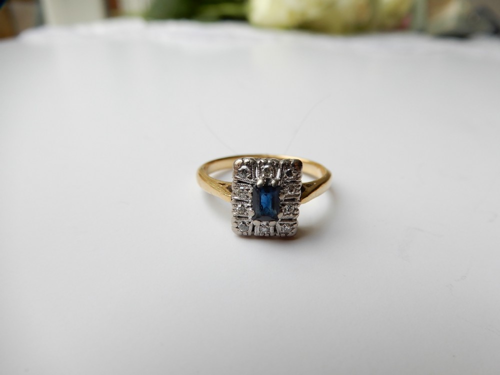 An Art Deco ring featuring a central rectangular blue sapphire surrounded by a geometric halo of small diamonds set in platinum atop an 18ct yellow gold band. View IV