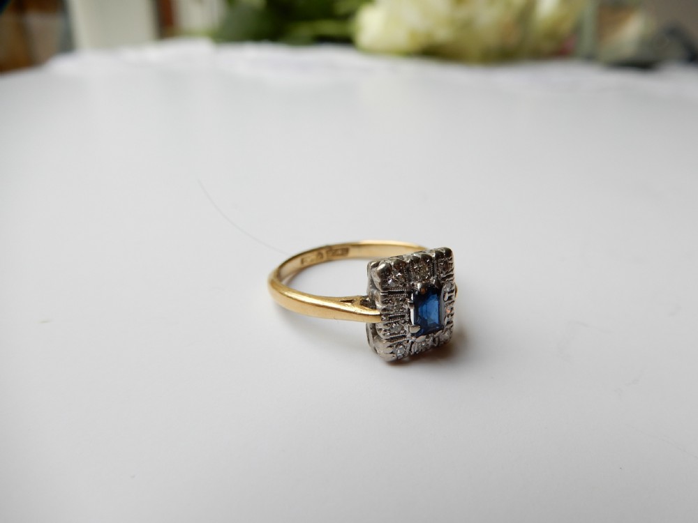 An Art Deco ring featuring a central rectangular blue sapphire surrounded by a geometric halo of small diamonds set in platinum atop an 18ct yellow gold band. View V