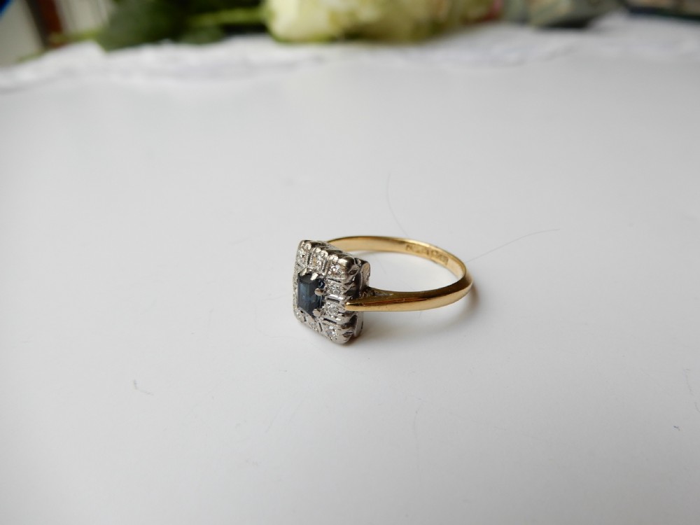 An Art Deco ring featuring a central rectangular blue sapphire surrounded by a geometric halo of small diamonds set in platinum atop an 18ct yellow gold band. View VI