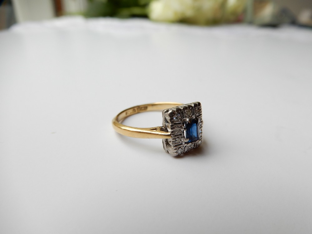 An Art Deco ring featuring a central rectangular blue sapphire surrounded by a geometric halo of small diamonds set in platinum atop an 18ct yellow gold band. View VII