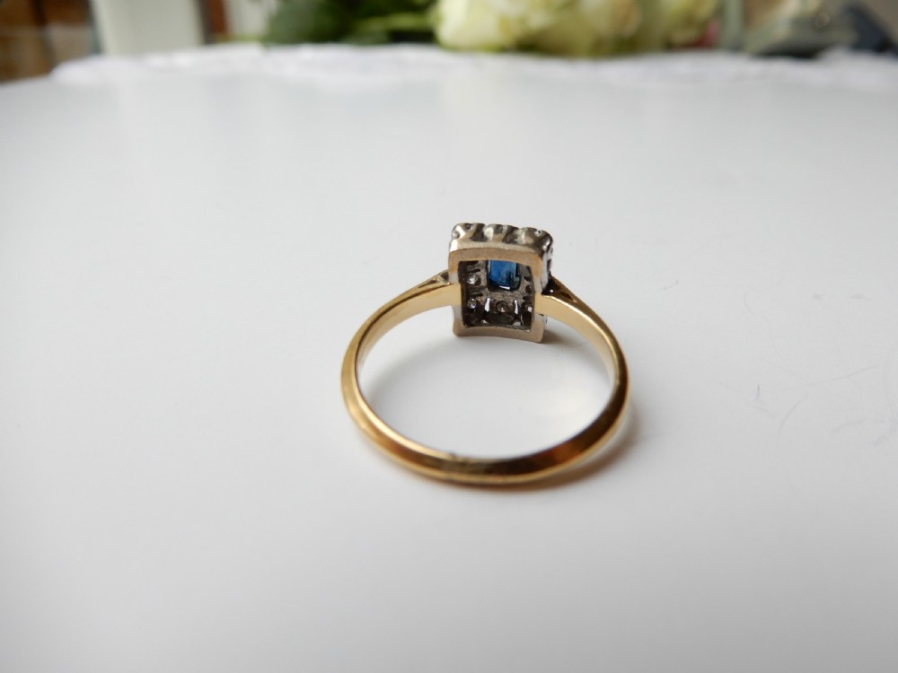 An Art Deco ring featuring a central rectangular blue sapphire surrounded by a geometric halo of small diamonds set in platinum atop an 18ct yellow gold band. View IX