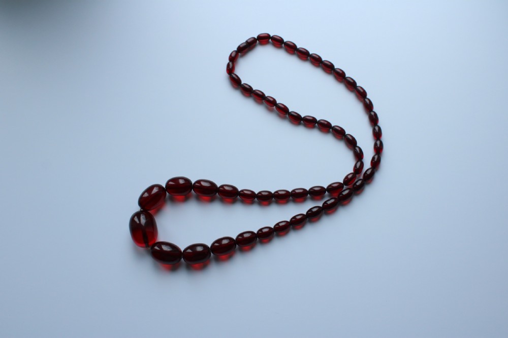 A continuous graduated strand of translucent cherry-red oval Bakelite beads displayed against a white background. View III
