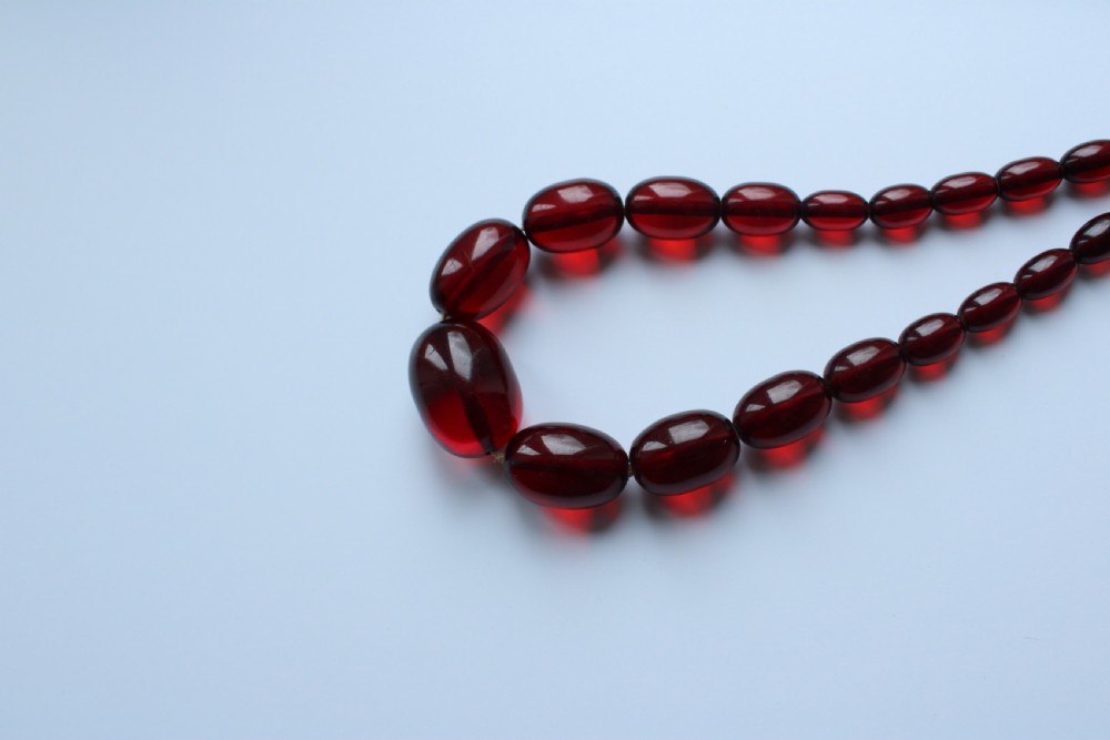 A continuous graduated strand of translucent cherry-red oval Bakelite beads displayed against a white background. View IV