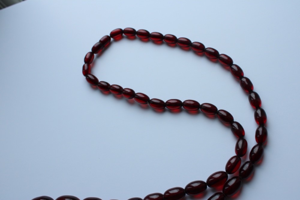 A continuous graduated strand of translucent cherry-red oval Bakelite beads displayed against a white background. View VI
