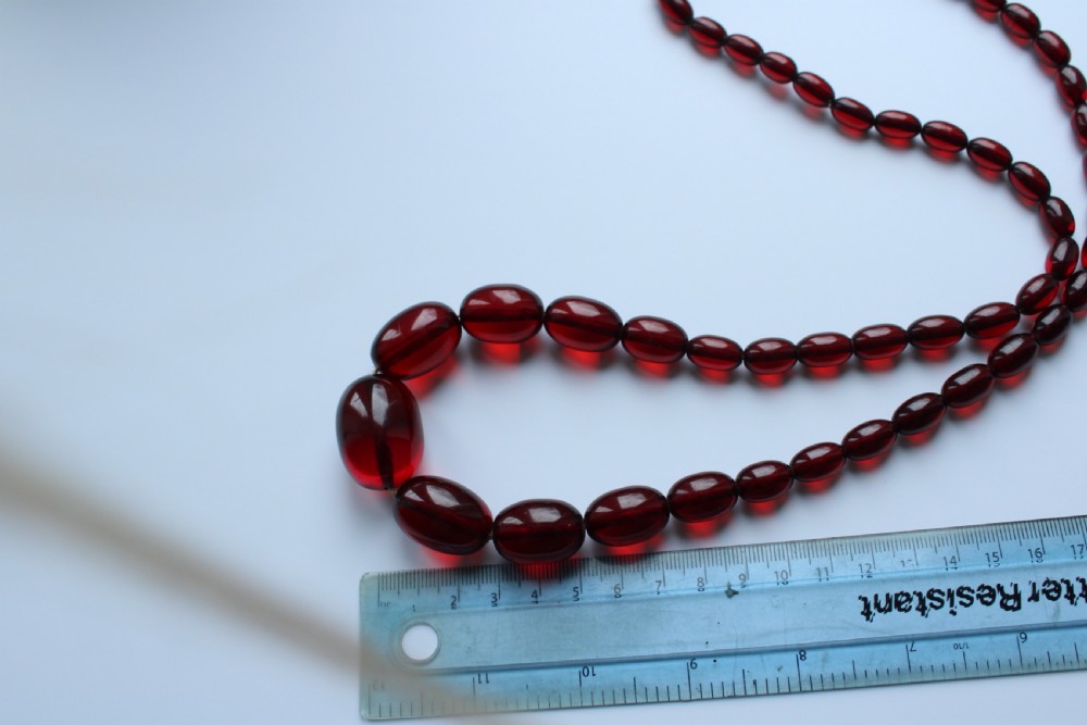 A continuous graduated strand of translucent cherry-red oval Bakelite beads displayed against a white background. View VII
