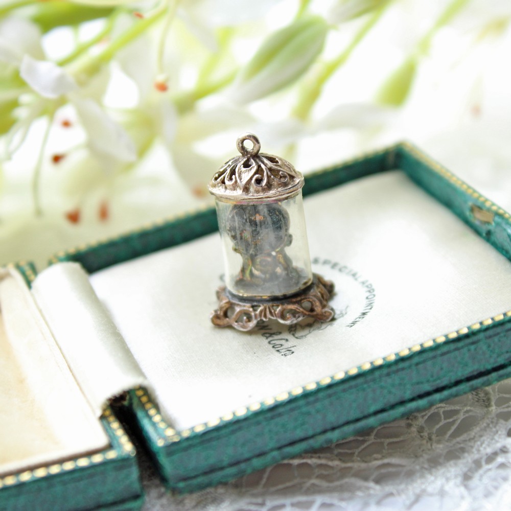 A miniature sterling silver charm featuring a mantle clock encased within a clear glass dome, decorated with ornate filigree detailed top and bottom.