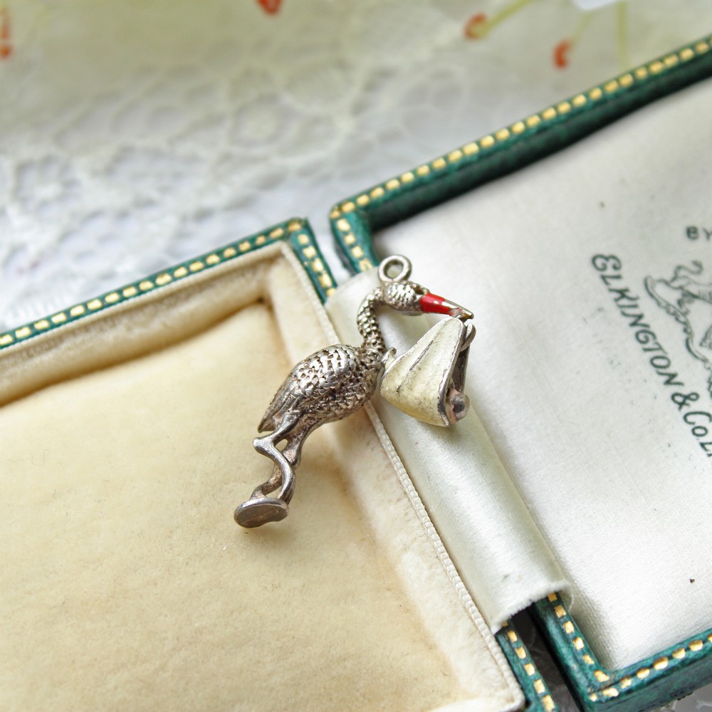A small sterling silver charm depicting a stork carrying a swaddled baby bundle with red and cream enamel accents.