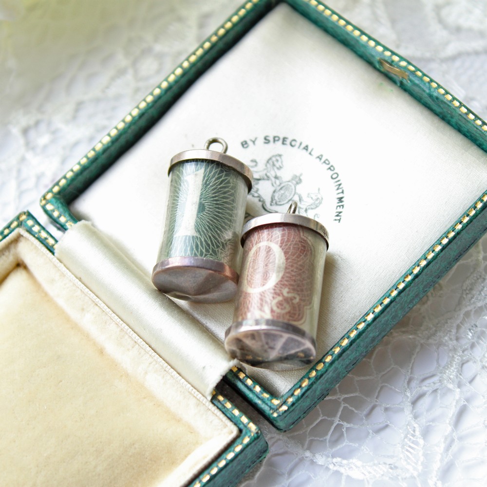 A pair of cylindrical sterling silver charms enclosing a rolled green one pound note and a red ten shilling note.
