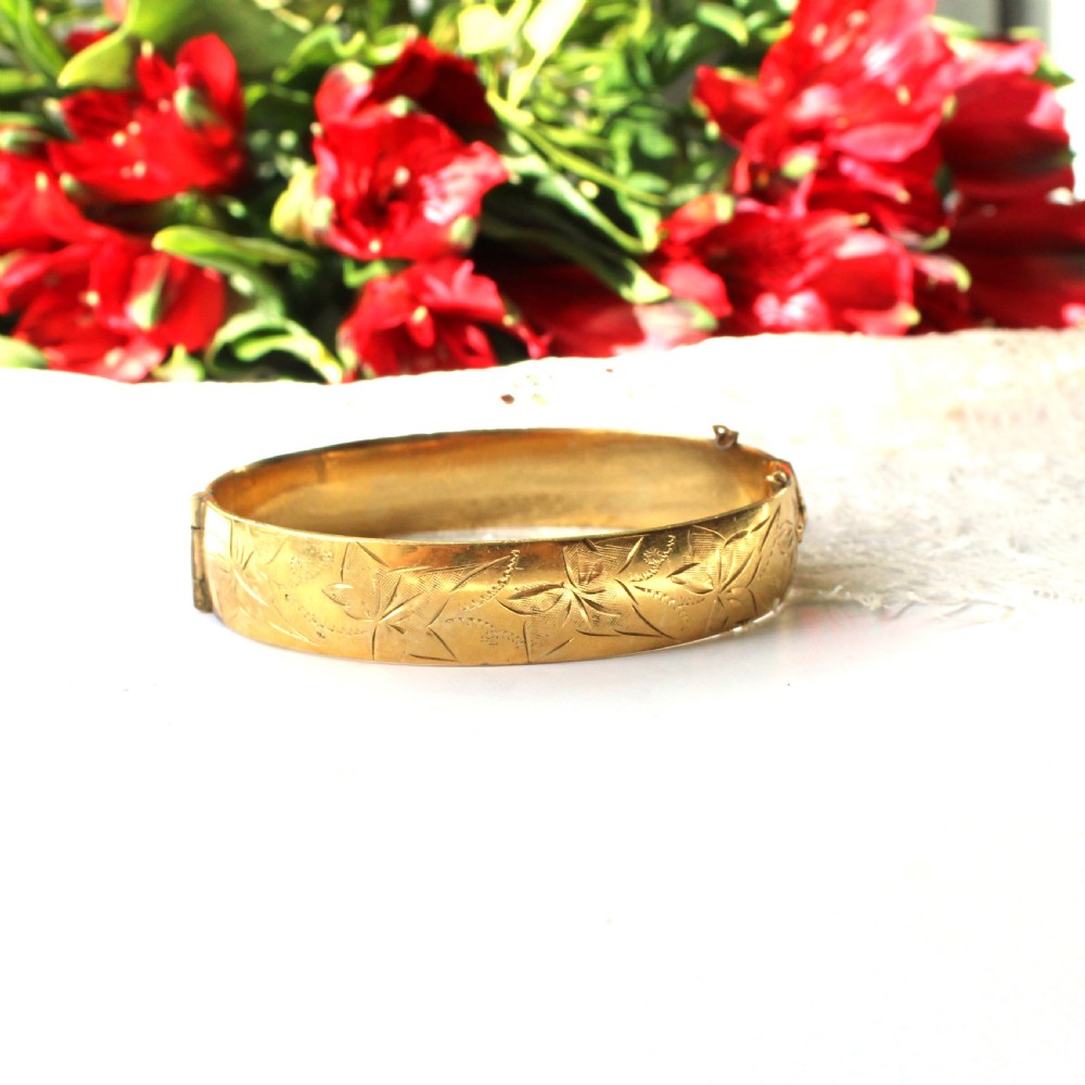 A vintage gold-toned hinged bangle bracelet featuring etched floral and leaf motifs around the outer circumference.