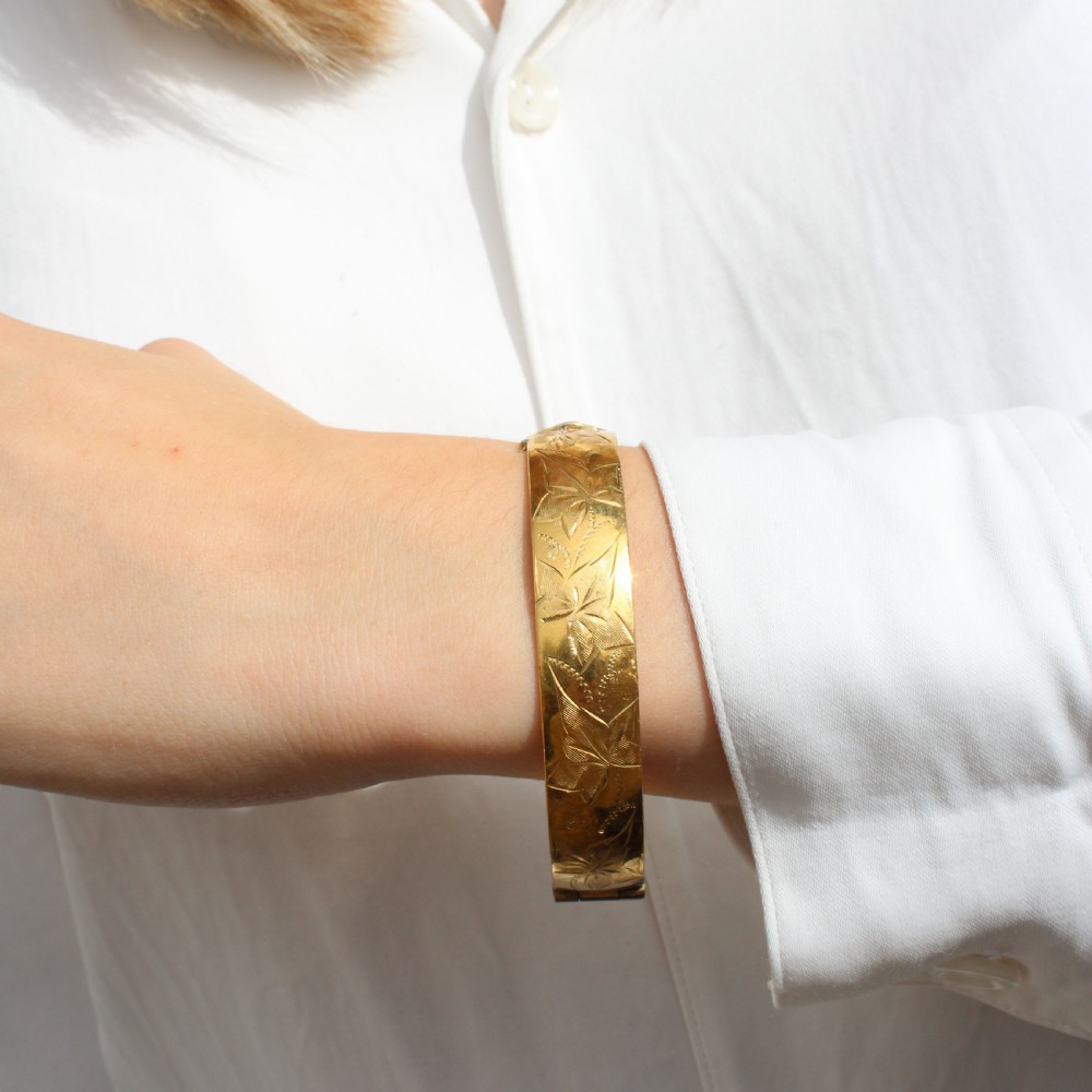 A vintage gold-toned hinged bangle bracelet featuring etched floral and leaf motifs around the outer circumference. View VIII