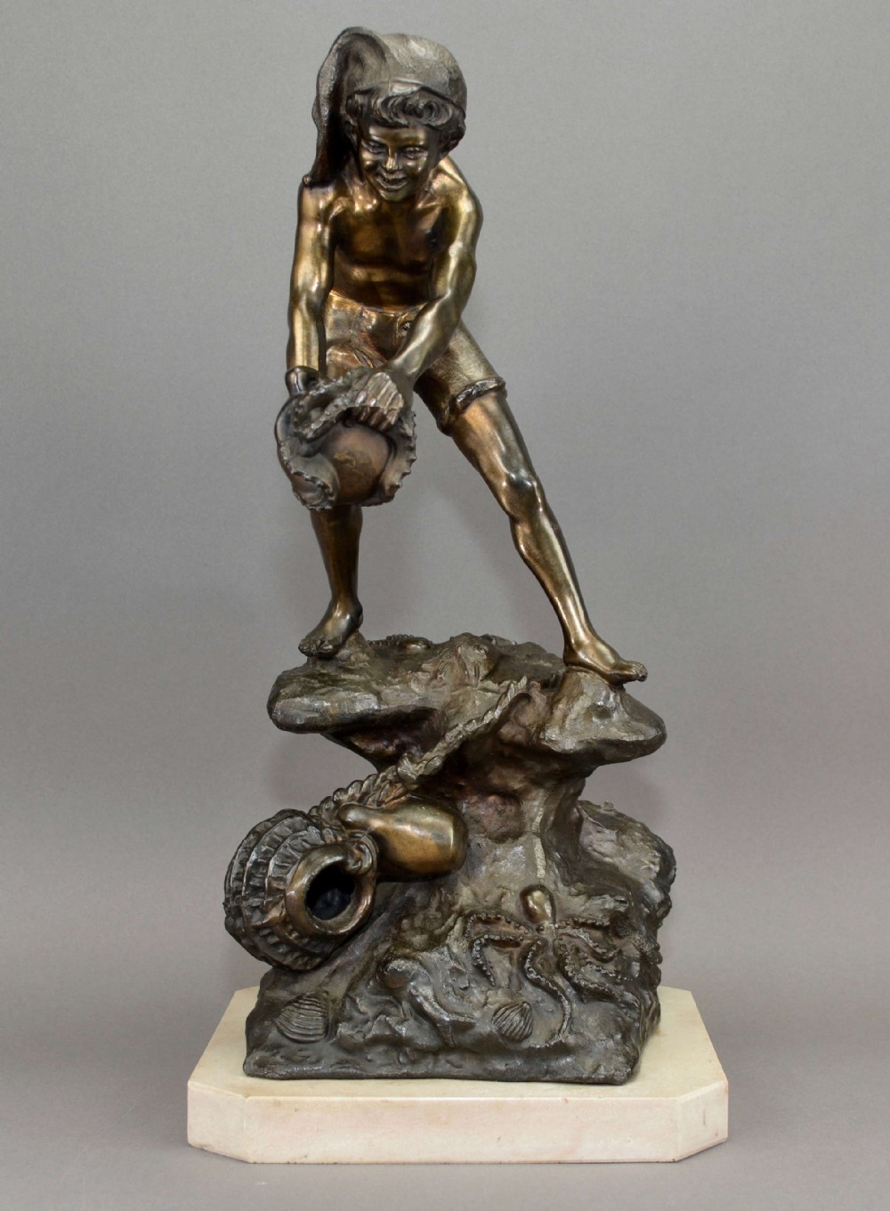 A dark bronze figure depicting a young fisher boy wearing a cap and shorts, standing on a rocky base decorated with sea life and mounted on a marble plinth.