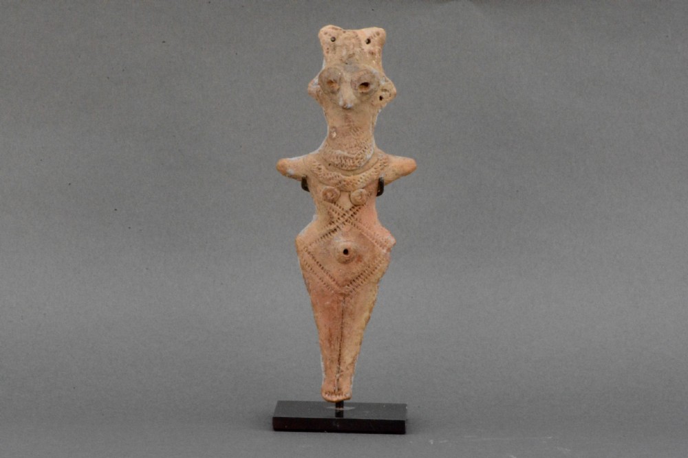 An ancient Syro-Hittite terracotta figure of the goddess Astarte featuring a bird-like face and incised geometric body markings mounted on a black stand.