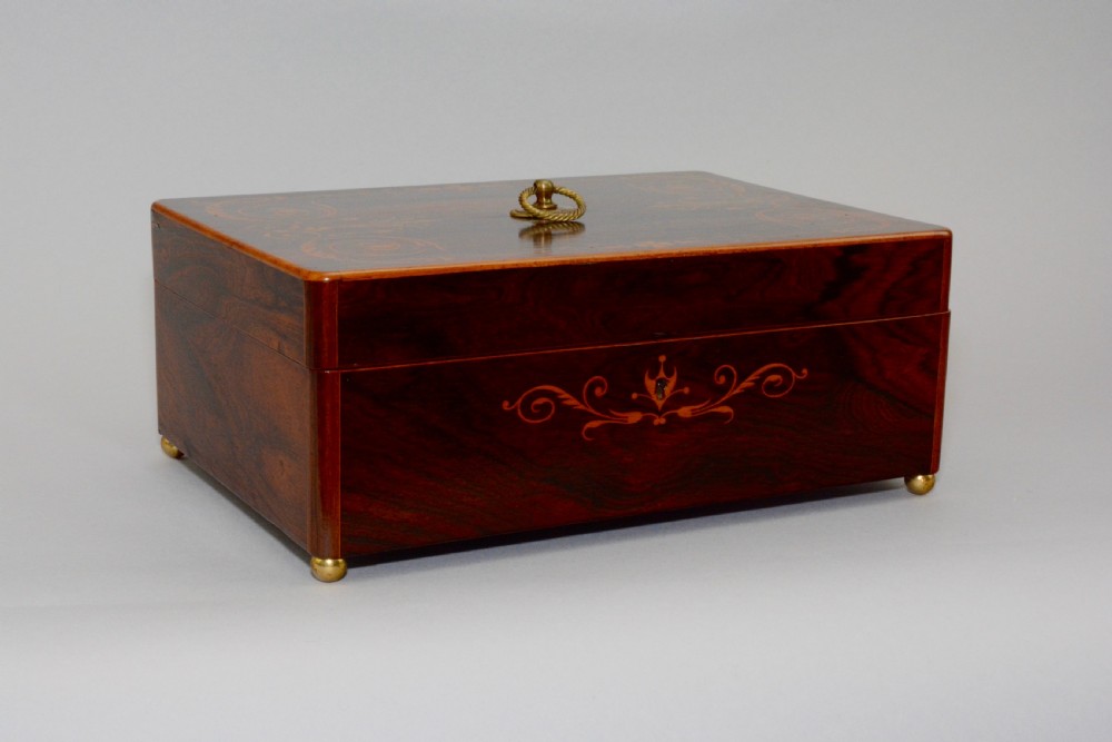 A rectangular dark reddish-brown rosewood box featuring fine wood marquetry around the keyhole, a top brass ring handle, and brass ball feet.