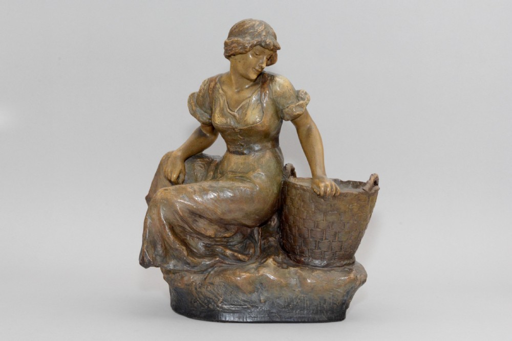 An Art Nouveau terracotta sculpture depicting a seated country maiden in draped clothing beside a woven basket.